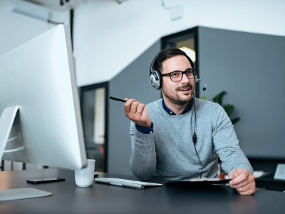 A man in a gray sweater and headphones points to his desktop monitor.